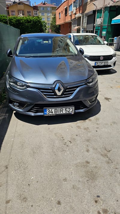 A dark grey Renault car is parked on a paved street in Ataşehir, Turkey. The car's front license plate reads "34 BIR 523 TR". Behind the Renault, a white Fiat car is also parked, its license plate reading "34 CAT 622". Buildings with apartment windows and balconies line the street. In the background, there are trees and other structures typical of an urban environment. The weather appears sunny.