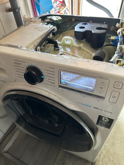 The image shows a white Samsung VRTplus 12.0kg washing machine, partially disassembled, in what appears to be a residential setting. The washing machine is in an upright position. The top panel of the washing machine is open, revealing internal components. The control panel, with a digital display and physical buttons, is visible on the front. The display shows "Шерсть 50 мин." which translates to "Wool 50 min." and indicates the selected wash cycle and time. The machine has a black front-loading door. The floor appears to be tiled. The background includes pipes and wires, suggesting the machine is undergoing maintenance or repair. No people are present in the image. The location is identified as Nakhabino, Russia.