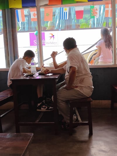 A young boy in a white t-shirt and black and white sneakers is sitting at a dark wooden table in a cafe in Tharpu, Nepal, with a bottle of water and a plate of food in front of him. He is looking down at his plate. An adult male in a light-colored patterned shirt and light-colored pants is sitting across from the boy, with his arm resting on the table. The cafe has a large window with flags hanging from the ceiling. Outside the window, a bus is visible with "Swift Holidays" printed on the side. A woman in a pink t-shirt is visible through the bus window.