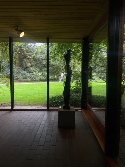 The scene depicts an interior architectural space in Humlebæk, Denmark, featuring a panoramic view of a lush exterior garden through a floor-to-ceiling glass wall. The interior floor is composed of dark reddish-brown brick tiles laid in a running bond pattern. Above, a light-toned slatted wooden ceiling is visible, from which a single, circular spotlight emits a warm yellow glow in the upper left quadrant.
Positioned near the center-right of the interior, directly before the glass wall, stands a tall, dark, abstract sculpture. The sculpture, possibly metallic or dark wood, rests on a grey rectangular plinth and exhibits an elongated, stylized form with curved lines. The expansive glass wall is framed by dark vertical mullions, with a dark wooden doorframe visible on the far right.
Beyond the glass, the exterior landscape features a meticulously maintained, vibrant green lawn in the immediate foreground, with dark green ivy or similar ground cover at the base of the windows. A winding, light-colored path traverses the mid-ground. The garden contains numerous mature trees, including both deciduous varieties with full green canopies and coniferous trees with blue-green needles. Several human figures are observed in the distance within the garden: two individuals, one appearing to be an adult in a white top and a smaller figure in light-colored attire, walk along the path towards the left. Additional indistinct figures are visible further back and to the right within the garden space.
