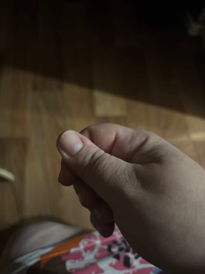 The media file shows a close-up of a person's hand, specifically the thumb and index finger, held in a pinching gesture. The hand is positioned over a wooden surface, likely a floor, with visible wood grain. The lighting is dim, creating shadows that obscure parts of the background. A portion of colorful fabric, possibly clothing or bedding with a pink and white pattern, is visible on the right side of the frame. The focus is on the details of the skin and fingernail of the person's thumb. There is no visible text. The overall impression is that of an intimate, everyday moment captured in low light. The location context provided is Княгинино, Russia, but this cannot be visually confirmed from the image itself.