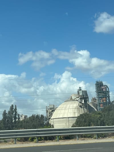The image shows an industrial area with a large, dome-shaped building and various industrial structures, including towers and interconnected piping, located next to a road. The sky is blue with scattered clouds. A guardrail separates the road from the industrial site. The image was taken in Ramla, Israel.