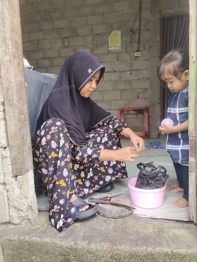A woman in a black hijab and a floral print dress, sitting on a concrete step outside a building, is working on a task with a plastic bag and what appears to be some sort of string or wire. She is facing away from the camera and is positioned to the left of the frame. A young child, wearing a blue checkered shirt, stands to the right of the frame, holding a light purple ball. The child is looking down at the woman. The background consists of a brick wall and a doorway. The setting appears to be in a residential area, possibly Cintadamai, Indonesia.