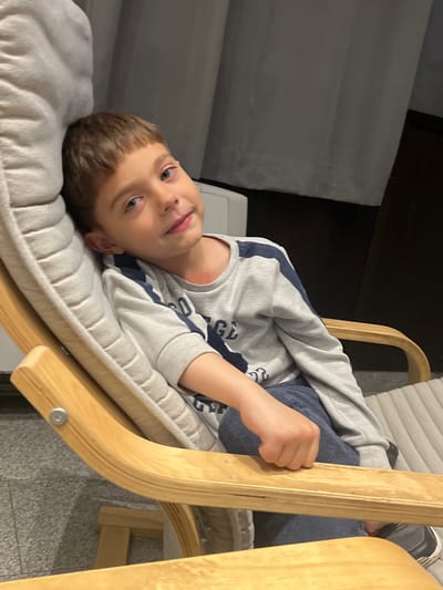 A young boy reclines in a light wood rocking chair with a gray cushioned seat and back. He wears a gray sweatshirt with blue accents and dark pants. His head rests on the seat cushion, and he looks at the camera. Part of a gray couch is visible behind the chair to the left, and a dark wood piece of furniture is above the chair. The photo is taken indoors. There is no information to suggest the photo was taken in Dedovo, Bulgaria.