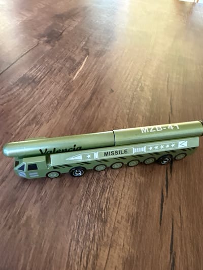A green toy missile launcher with a wheeled vehicle base sits on a wooden surface. The missile itself is cylindrical with the text "MZB-41" printed vertically on its side. Below this, "Valencia" is written in a stylized font. The wheeled vehicle base has multiple sets of wheels and what appears to be a control panel with lights or indicators. There is also text on the vehicle's side, including "MISSILE" and several star symbols within a rectangular box. The overall scene suggests a close-up shot of a toy, likely for play or display. The lighting is bright, indicating it's daytime. The wooden surface suggests an indoor setting, possibly a table or desk. No people are visible.