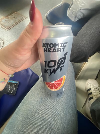 A person is holding a silver can of "ATOMIC HEART 100 KWT" energy drink. The can has a grapefruit slice graphic and text. The person's left hand, with long, dark red nails, is visible, wearing a red string bracelet with beads and a small colorful flower tattoo on the wrist. A black wallet with a rhinestone star is also visible. The person is wearing light blue jeans and a black shoe with white stripes. The background shows a dark blue seat, a metal grate on the floor, and a gray bag. The scene appears to be inside a mode of transport, possibly a train or bus. There is no specific information in the image to indicate a location in Aprelevka, Russia.
