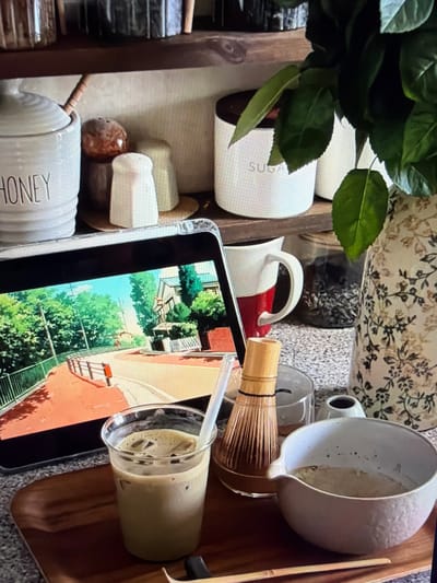 An overhead view captured from a slightly elevated angle shows a collection of items arranged on a wooden tray or caddy, resting on a speckled countertop. The scene is set indoors, contextually in Ros Na Rí, Ireland.

In the upper left quadrant, a cluster of dark green leafy foliage partially obscures a white ceramic jar with the word "SUGAR" visible on its side. To the right of the sugar jar, a white mug with a red base and handle is visible. Further right, a light-colored, patterned fabric or ceramic piece is in the background.

A white, spouted ceramic pitcher or chawan, containing a light brown residue, is positioned towards the upper right. Adjacent to it, a bamboo matcha whisk (chasen) stands upright. Below the whisk, a clear plastic cup holds an iced, frothy, light brown drink (likely a matcha latte or coffee) with ice cubes and a transparent straw.

Dominating the center of the frame is a tablet or smartphone, its screen displaying a vibrant animated image of green trees, blue sky, and buildings with red roofs, suggesting a pleasant outdoor scene.

In the lower left, another white ceramic jar is visible, clearly labeled "HONEY" with a wooden honey dipper resting inside. Various smaller ceramic containers and objects are scattered around these main items.

The activity depicted suggests a moment of leisure and drink preparation or enjoyment, possibly involving matcha, while consuming media on a digital device. The overall atmosphere is one of domestic comfort and relaxation. No people are visible in the frame.