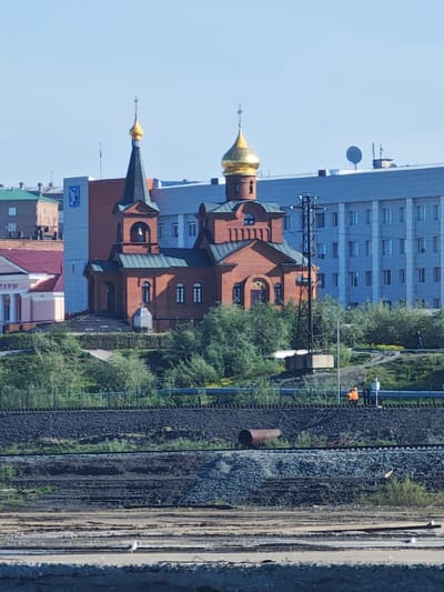 This is a daytime, outdoor, long-shot photograph taken in Dudinka, Russia. The scene depicts an Orthodox church with a prominent gold dome and a dark green steeple, constructed from red brick. The church is situated on a grassy hill, overlooking a body of water in the foreground. Behind the church, a large, light blue, multi-story building dominates the background, with many windows. To the left of the church, a smaller pink building with a burgundy roof is partially visible.

In the middle ground, a network of train tracks runs horizontally across the image, with piles of dark, coarse material and some scattered greenery alongside them. A rusty barrel is visible on the tracks. On a rise above the tracks and in front of the large blue building, two individuals in orange vests can be seen, appearing to be workers. Further to the right, a single person in dark clothing stands near the blue building. The sky is a clear, bright blue, suggesting fair weather and likely midday. The overall impression is one of an urban environment with industrial elements and a prominent religious structure.