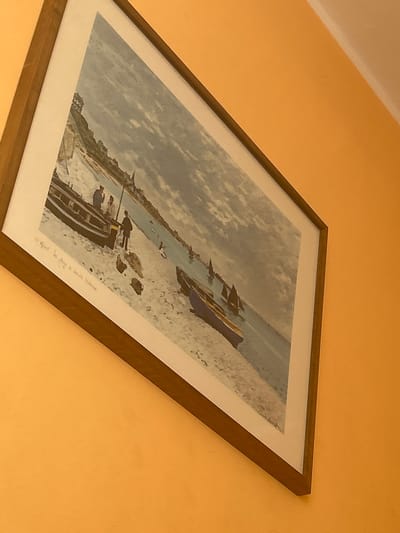 A framed artwork is mounted on a textured, bright orange wall. The artwork depicts a beach scene with a body of water and several boats. In the foreground, there are two boats on the sand, with three figures standing near one of them. The figures appear to be men, one in a dark outfit and the other two in lighter clothing. The water is a muted blue-grey, and the sky is cloudy. Further back in the painting, there are more boats and buildings along a coastline. A handwritten inscription in cursive is visible at the bottom left of the artwork. The image is taken from a low angle, looking upwards at the artwork. The overall lighting suggests an indoor environment, possibly a home or gallery, given the wall color and the way the artwork is displayed.