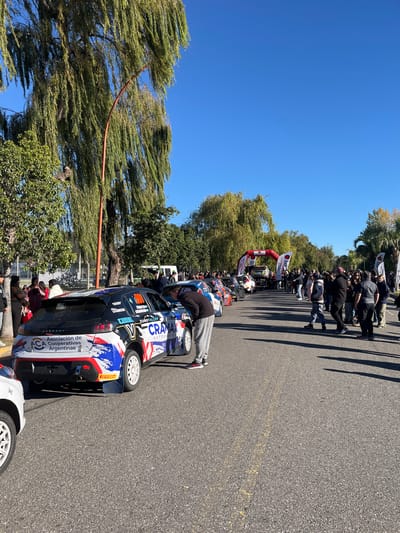 The image shows a street in Villa Carlos Paz, Argentina, lined with parked rally cars and spectators. Several people stand around a dark-colored rally car with sponsor logos and the words "Asociación de Cooperativas Argentinas." Spectators are positioned along the road, some under trees. An inflatable archway spans the road further down the street. The sky is clear and blue.