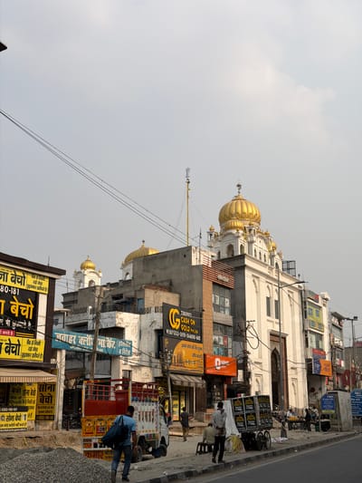 The image depicts a street scene in Panipat, India, on a bright, overcast day. The dominant structure is a Sikh temple, identifiable by its white exterior and golden domes, a prominent feature against the pale sky. The temple is flanked by a row of commercial buildings, their facades adorned with numerous signs and advertisements in various languages, including Hindi and English.
The street level is bustling with activity. Several individuals are visible, some engaged in conversation or looking at their phones, while others appear to be conducting business. A man in a blue shirt and jeans is seen walking with a bag slung over his shoulder, navigating the crowded sidewalk. A truck, possibly used for transportation or delivery, is parked on the side of the road, its back filled with items. Carts and bicycles are also present, suggesting common modes of transport and commerce in the area.
The advertisements plastered on the buildings and displayed on signs offer a glimpse into local businesses and services. Some signs promote gold buying and selling, with prominent text like "TIRUPATI CASH ON GOLD Pvt. Ltd." and "WE BUY GOLD SILVER-DIAMOND-BARS." Other signs are for hotels and markets, indicating a commercial hub. The overall impression is one of a vibrant, busy urban environment with a strong religious presence. The time of day appears to be daytime, given the bright but diffused light.
