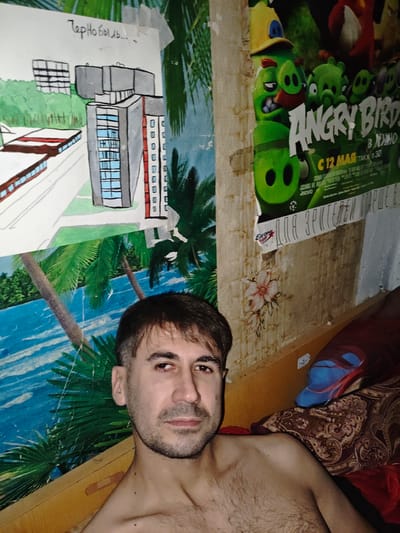 A man is captured in a close-up, chest-up selfie, positioned in the lower-right quadrant of the frame. He has short, dark brown hair, a receding hairline, and a beard and mustache. His skin appears to be slightly flushed, and he has a neutral expression. He is shirtless, revealing a hairy chest.
The background is an eclectic collage of posters and drawings. On the left, a colorful painting depicts a tropical scene with palm trees and a body of water, overlaid with a simplified drawing of modern buildings, possibly meant to represent a city or infrastructure project. Text above this drawing in Cyrillic reads "Чернобыль..." ("Chernobyl...").
To the right of the painting, a large poster for "Angry Birds" is visible, featuring cartoon characters from the film. Below this poster, a strip of wallpaper with a floral pattern is peeling away. A blue and red slipper rests on a patterned comforter or blanket in the upper right corner. The overall environment suggests a room with a somewhat cluttered and personalized decor. The image is dimly lit, casting shadows and contributing to a slightly gritty atmosphere.
While the image is consistent with an indoor residential setting, it is not possible to definitively ascertain the precise location within Akhmetovskaya, Russia, based solely on the visual information provided.