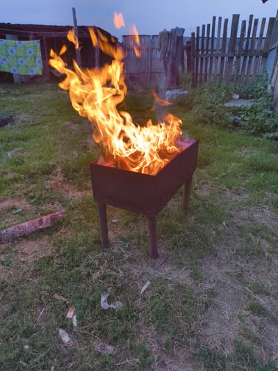 A fire burns brightly in a rectangular metal brazier with four legs, set on a grassy lawn. The flames are tall and vibrant, casting a warm glow. Behind the brazier, a rustic wooden fence encloses the area. To the left of the brazier, a piece of wood lies on the grass. In the background, a building with a dark roof and a clothesline with a green and white patterned cloth are visible. The sky is a dusky blue, suggesting it is either dawn or dusk. The overall scene evokes a sense of outdoor leisure or a quiet evening.