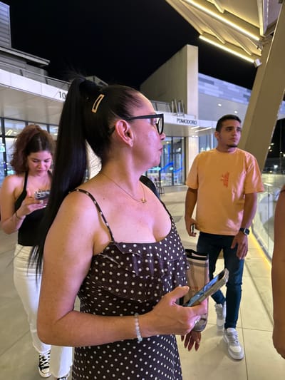 The image depicts a group of people in an urban outdoor setting at night. The primary subjects are three individuals. In the foreground, a woman with dark hair pulled back in a ponytail, wearing glasses and a brown polka dot dress, is looking upwards. Her left arm, adorned with a light green beaded bracelet, is visible, as is her right hand holding a smartphone. Behind her, another woman with reddish-brown hair is also holding a smartphone and appears to be taking a selfie. In the background, a man in an orange t-shirt and dark jeans is standing, holding his phone and looking to the side. He has a watch on his left wrist.
The setting appears to be a modern urban environment with sleek architectural elements and bright lighting. There are white beams and illuminated panels visible, suggesting a contemporary building or public space. A sign with the text "POMODORO" is visible in the background. The lighting suggests it is nighttime. The mood seems casual and relaxed, with people engaged with their phones and in conversation or observation. The overall scene suggests a social gathering or a moment captured while out and about in a city at night. The location is identified as Alcorcón, Spain.