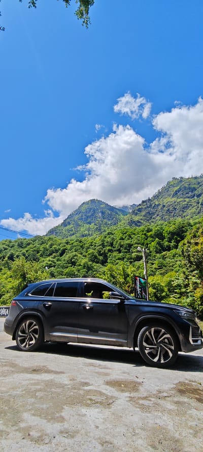 A dark-colored SUV, likely black or dark grey, is parked on a light-colored paved surface by the side of a road, set against a stunning backdrop of lush green, forested mountains. The car features tinted windows, silver trim along its windows and lower body, and distinctive alloy wheels with a swirling, multi-spoke design. A person's silhouette is faintly visible through the driver's side window.

The mountains are densely covered in vibrant green trees, appearing steep and rugged, with some peaks touching the bright blue sky. Numerous white, fluffy clouds are scattered across the sky, particularly concentrated around the mountain peaks on the right. Bright sunlight illuminates the scene, suggesting a clear day, likely morning or early afternoon, with a subtle sun flare visible in the upper left corner of the sky. A few green tree branches hang into the frame from the very top left.

Near the front of the car, a tall street light pole is visible, and beneath it, an electronic sign or screen displays a colorful, somewhat glitched pattern. The paved ground beneath the car shows some wear and patches of dampness. On the far left, partially obscured by the car's front door, a white sign with Cyrillic letters is visible, showing "ЙКА," which could be part of a word like "СТОЙКА" (meaning "stand" or "counter"). This scenic moment is captured in a mountainous region, specifically Bzypta, Abkhazia, highlighting a travel stop amidst natural beauty.