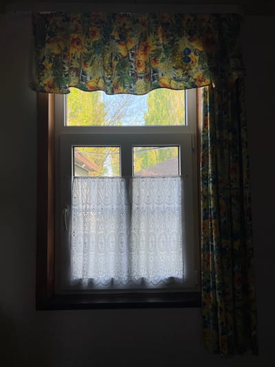 The image depicts a window in a dimly lit room. The window is divided into two panes, with a view to the outside visible through the left pane and a white lace curtain covering the right pane. The outside view shows green trees and a glimpse of a rooftop under a blue sky, suggesting it is daytime. A decorative, floral-patterned curtain is pulled back on the left side of the window. A wooden window sill is visible. The surroundings are dark, with the room's walls barely discernible. There is no direct evidence to confirm the location in the city of Samarskoye, Russia.
