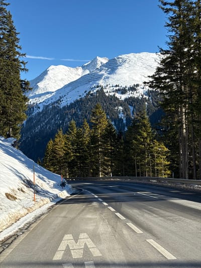 The image presents a winter alpine scene depicting a paved road in St. Christoph, Austria, under a clear blue sky. The asphalt road, featuring white dashed lane markings and a large white "M" symbol painted on its surface in the foreground, curves slightly to the right as it recedes into the distance. To the immediate left of the road, a snow-covered bank rises, punctuated by two slender poles with orange tops. A metal guardrail runs along the right edge of the road. Dense evergreen conifer trees line both sides of the road, extending up the lower slopes of the mountains. In the background, large snow-covered mountains dominate the landscape, exhibiting distinct parallel terracing or patterns on their white slopes, possibly indicative of avalanche control measures or groomed ski terrain. No individuals or vehicles are visible within the frame.