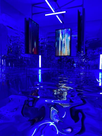 The image depicts an immersive art installation with a dominant blue color scheme. The environment features highly reflective, rippling surfaces that suggest water or a similar liquid medium covering the floor. Tall, narrow vertical light fixtures emit a bright white-blue light, and horizontal fluorescent tubes are visible near the ceiling, casting a cool blue glow. Two large, vertically oriented digital screens are mounted on stands. The screen on the left displays abstract, colorful imagery with hints of red and white, while the screen on the right shows a more geometric, abstract scene with blues, whites, and some lighter tones. The walls also appear to be made of a shiny, reflective material, amplifying the light and creating a disorienting, futuristic atmosphere. There are no people visible in the scene. The specific location in Jinniu District, China, cannot be determined from the visual information alone.
