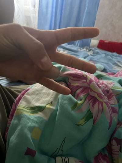The image shows a person's hand making a peace sign in the foreground. The hand is positioned over a brightly colored fabric with a floral pattern, predominantly teal, pink, and green. In the background, a blue curtain is visible, and a bed with blue bedding can be seen. The overall scene suggests an indoor setting, possibly a bedroom. There is no information in the image that can identify its specific location within the city of Узяр, Russia.