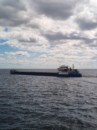 A wide shot shows a long blue and white barge moving on water. The barge has a control cabin on the right side with a visible name in Cyrillic script, suggesting the location may be Russia. The water is dark and choppy. Above, the sky is filled with white and gray clouds with patches of blue sky peeking through. There are no people visible in the image. The photograph appears to be taken during the daytime under overcast conditions. No specific details indicating Mologa, Russia are present within the imagery.