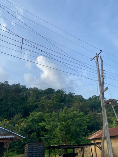 The image, taken in Igarra, Nigeria, presents a daytime scene dominated by a tree-covered hill under a partly cloudy sky. Numerous dark electrical power lines traverse the upper portion of the frame. A wooden utility pole, visibly split at its upper end, stands on the right side, supporting multiple insulators and the power lines. A street light fixture is attached to this pole, below which a smaller structure holds a solar panel. In the lower-left, a section of a building with a corrugated metal roof is visible. Further right in the foreground, a dark, rectangular wooden structure is present. Towards the lower-right, a building with a reddish-brown corrugated metal roof and a light-colored wall is partially obscured by the utility pole and surrounding vegetation. The midground and background consist of a densely forested hill or mountain featuring visible rocky outcrops. No individuals are depicted, and no specific actions are in progress.