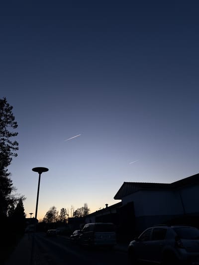 The image shows a street view in Lauchhammer, Germany, at twilight. The sky is a gradient from dark blue to light blue near the horizon. Two contrails from airplanes are visible in the sky. On the right side of the frame is a building and parked cars. A streetlight and trees are silhouetted in the foreground.