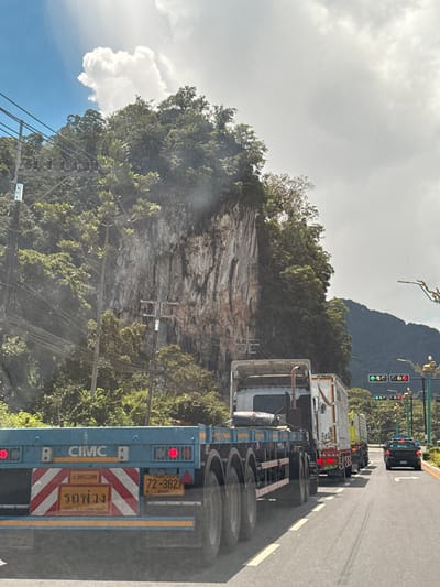 The image shows a road scene in Phang-nga, Thailand, with a large limestone karst mountain covered in lush green vegetation dominating the background. A blue flatbed trailer truck with "CIMC" branding is in the foreground, displaying red taillights and a red and white reflective chevron pattern on its rear. A Thai license plate reading "72-3627" is visible on the trailer, along with Thai text that appears to be "รถพ่วง" (rot phuong), meaning "trailer."

Following the trailer truck are several other trucks, including a white truck with a box trailer. A black pickup truck is in the right lane of the road, approaching a traffic light. The traffic light displays green arrows pointing left and right for the top row, and a red light for the bottom row. Streetlights and power lines are also visible, crisscrossing the scene. The sky is partly cloudy, with bright white clouds against a blue sky. The overall impression is a typical road scene in a tropical, mountainous region of Thailand.
