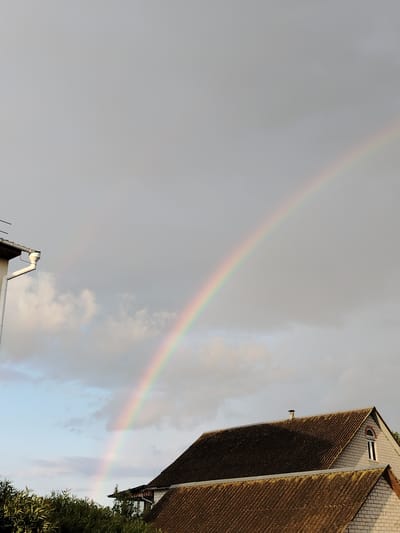 A rainbow arcs across a cloudy sky over a residential area. In the foreground, two roofs are visible, one with dark shingles and the other with a corrugated, weathered appearance. A portion of a white building with a downspout is on the left. The sky is a mix of gray and lighter clouds, with patches of blue visible. The scene appears to be set in an outdoor environment, possibly during or after a rain shower, in the city of Rudzensk, Belarus. There are no people or animals visible in the image.