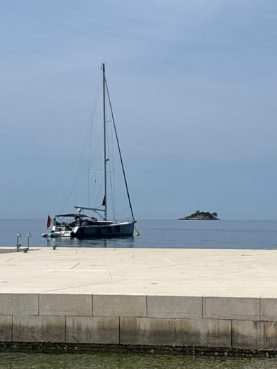 A horizontal outdoor shot shows a sailboat docked on a concrete pier near Grad Rovinj, Croatia. The yacht is dark grey, with a tall mast and several antennas and is decorated with flags. It is moored alongside the pier, which is made of large, rectangular concrete blocks. The pier is bordered by a thin strip of water and rock. The sea is a light blue, with a small, grassy island visible in the distance. The sky is a clear, light blue.