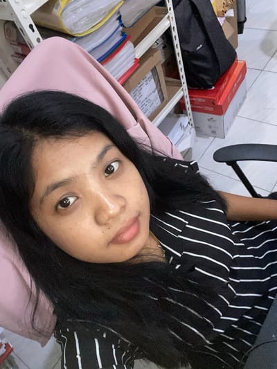A person with dark hair and light skin is lying down, facing the camera. They are wearing a black and white striped shirt. Behind them is a light pink surface. To the left, there is a shelf with various objects including a black bag, red and white boxes, folders, and other items. The background suggests an indoor location, possibly an office or storage area. The picture may have been taken indoors, possibly in Negara, Indonesia based on the user's request.