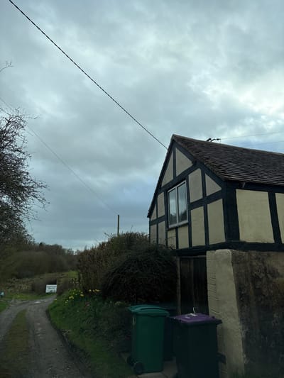The image depicts the exterior of a half-timbered house in Shelve, United Kingdom, under a cloudy sky. The house has a distinctive architectural style with dark timber framing contrasting against lighter infill panels. A single window is visible on the side of the house. Two wheelie bins, one purple and one green, are positioned adjacent to the house. A hedge is growing near the house. The scene includes a dirt path and a sign reading "SMARTSTONE HOLLOW". A power line stretches across the sky. Bare trees and bushes are visible in the background.