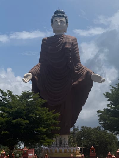 A low-angle, full-shot captures a monumental, standing Buddha statue against a partly cloudy sky in Na Nuea, Thailand. The statue, draped in a dark brown robe, has its arms extended outwards, palms facing forward. The face of the Buddha is rendered in a pale, serene expression. In the foreground, lush green trees partially obscure the base of the statue, which appears to be undergoing construction or renovation, indicated by scaffolding and supporting structures. Decorative red and orange architectural elements are visible at the very bottom of the frame. The overall impression is one of grandeur and spiritual significance.