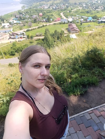 A young woman with light brown hair tied back is taking a selfie outdoors. She is wearing a dark red ribbed tank top with a black bra underneath and a long pendant necklace. Sunglasses are hooked onto her top. She has freckles on her face and is looking towards the camera with a neutral expression. Behind her, the background shows a view of a village with houses, roads, and green vegetation. A body of water, possibly a river or lake, is visible in the distance, along with rolling hills covered in trees and foliage. The foreground shows a path paved with colored bricks and overgrown grass. The scene appears to be in a rural or semi-rural setting, consistent with the outskirts of Solnechnaya Polyana, Russia, given the landscape and architecture.
