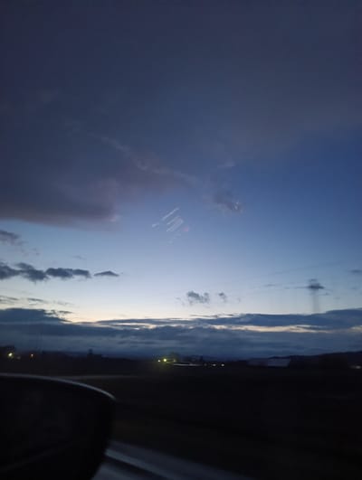 The photograph depicts a view from inside a vehicle at dusk or dawn. The sky is a blend of dark blue and gray, indicating low light conditions. Clouds of varying shapes and sizes are visible. In the middle distance, there are hints of a landscape, possibly rural, with dark silhouettes of buildings and trees. Electrical wires stretch across the sky. The foreground is dominated by the interior of the car, including a side mirror and the dashboard. The location is unknown, therefore, I can't assert that this image was taken in Gory, Russia. No people are visible.