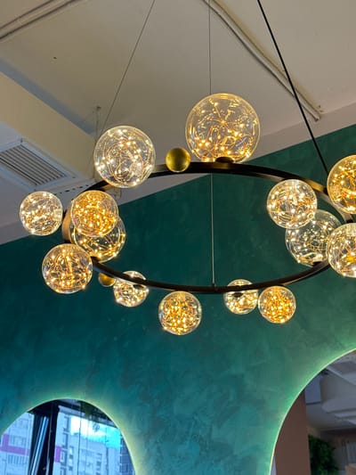 This is a medium shot, eye-level photograph of a modern chandelier with multiple glass orbs filled with fairy lights. The chandelier is suspended from thin black wires against a backdrop of a teal-colored, abstract ceiling structure and a white wall with exposed conduit.
The chandelier features a large, dark, circular frame from which numerous glass spheres hang. Each sphere contains a dense network of small, warm white LED lights that give them a glowing, festive appearance. The orbs vary in size, with some larger and others smaller, and they are arranged both within and around the circular frame.
The teal ceiling structure has a textured, organic shape, creating a contemporary and artistic feel. The white wall on the left side appears to be part of an interior space, with a visible air vent or grate. The lighting is warm and inviting, suggesting an indoor setting, possibly a restaurant, cafe, or modern living space. There is no visible text in the image. The time of day and weather are not discernible from the image.
