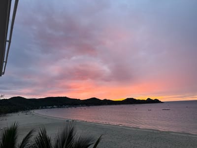 The image captures a serene sunset over a beach in Hòa Hiệp, Vietnam. A crescent-shaped beach with fine sand stretches along the coastline, dotted with a few scattered beach chairs and small boats. The calm sea reflects the vibrant colors of the sky, transitioning from a pale pink and purple near the horizon to a softer lavender higher up. Lush green hills form a backdrop to the beach, with a small village nestled along the shore, its buildings visible as white specks. Several boats are moored in the shallow waters. In the foreground, the dark silhouettes of palm fronds add a tropical touch to the scene. The sky is filled with wispy clouds, painted with hues of orange, pink, and purple, indicating the sun has recently set or is about to rise. The overall impression is one of tranquility and natural beauty.