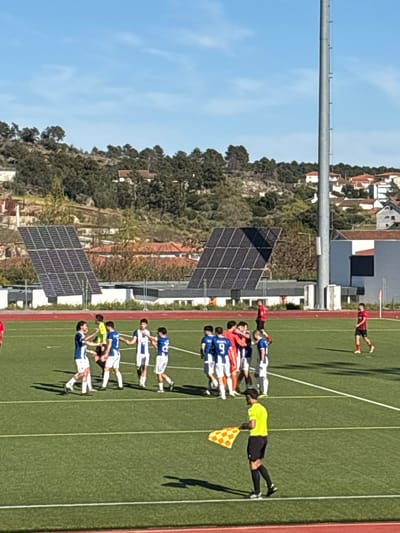 A soccer game takes place in Valpaços, Portugal. A group of players in blue and white uniforms are lined up on the green field. One man in a yellow shirt and black shorts, holding a checkered flag, stands next to the players. Another individual in red lays on the field in the background. Solar panels stand to the side of the field. Buildings and trees are visible in the background under a clear blue sky.