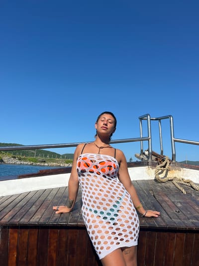 A person is seen relaxing on the wooden deck of a boat under a clear blue sky. The image is taken from a low angle, looking upwards, and is rotated 90 degrees clockwise, so the horizon appears vertical.
The individual, who appears to be a woman, is lying on her back, facing upwards towards the sky with her eyes closed, suggesting she is sunbathing or simply relaxing. She has short, dark hair. She is wearing a white, open-knit, short-sleeved dress or cover-up, which is semi-sheer and reveals an orange bikini top underneath, and a glimpse of what appears to be a dark green or teal bikini bottom on her lower body. A dark necklace is visible around her neck, and she wears a ring on her right hand. A tattoo is also visible on her right forearm.
The boat deck is made of dark wooden planks. Silver metal railings run along the side of the boat, separating the deck from the water. A thick, coiled rope is visible on the wooden deck near the railings. In the background, across the calm blue water, a green, hilly or mountainous landmass is visible. The sky is bright and cloudless, indicating a sunny day. The lighting suggests it is daytime, likely late morning or afternoon. The overall atmosphere is one of leisure and tranquility.
