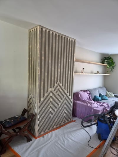 The image shows an indoor scene, likely a living room or a room undergoing renovation. A large, rectangular piece of furniture with a slatted wood design, featuring a geometric pattern, dominates the foreground. The wood appears to be a light, weathered gray. Behind this furniture, a grey sofa is visible, covered with a purple blanket and several decorative pillows in shades of blue and green. To the left of the sofa, a minimalist wooden shelf unit is mounted on the wall, displaying a potted plant and a few decorative items. In the background, a blue vacuum cleaner with hoses is partially visible, along with a metal ladder leaning against the wall, suggesting ongoing work or cleaning. The floor is covered with a white drop cloth, edged with orange tape, further indicating a renovation or protection scenario. The lighting suggests it is daytime, with light coming from the ceiling and possibly a window out of frame. No text is visible in the image. The scene conveys a sense of domestic space, possibly in a state of transition or preparation.
