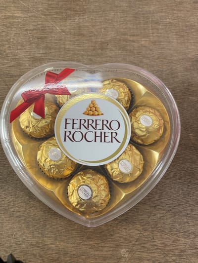 A heart-shaped clear plastic box containing Ferrero Rocher chocolates rests on a wooden surface. The box is tied with a red ribbon. The chocolates are individually wrapped in gold foil, and a circular label with the "Ferrero Rocher" logo is prominent in the center of the box. The wooden surface appears to be a tabletop or counter. The background shows a neutral wooden texture. This image does not contain information to determine its location in San Jose del Monte, Philippines.