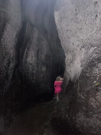 An individual wearing a bright pink dress is walking through a narrow canyon with wet, textured rock walls. The canyon floor appears to be covered in shallow water. The lighting is dim, suggesting an overcast day or the natural low light within the canyon. The scene is located in Bangli, Indonesia, likely at the Tukad Cepung Waterfall or a similar natural formation in the area. The individual is the sole subject, moving away from the camera deeper into the canyon.