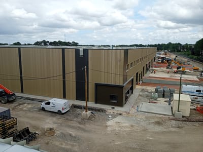 This image captures a large construction site in Mérignac, France, under an overcast sky. The primary subject is a substantial industrial or commercial building, largely completed, featuring a modern facade with alternating vertical panels of tan/light brown and dark grey/black. The flat roof is partially visible, showing some ventilation structures.
The foreground and midground are a bustling construction zone. The ground is a mix of dirt and gravel, with areas prepared for concrete pouring or paving. Numerous construction vehicles are present, including several yellow excavators, one with a large bucket, and a red-boomed telehandler on the far left. Several construction workers, clearly visible in their orange high-visibility vests, are engaged in various tasks across the site, seemingly preparing ground surfaces and managing materials.
Stacks of grey concrete blocks, pipes, and other construction materials are distributed across the site. A white utility van, prominently displaying the "TML" logo in blue and black, is parked on the dirt. Another white truck with a blue stripe is visible further down the site. In the distance, a line of green trees and a tall communication tower are visible against the cloudy sky. The scene depicts active development during daylight hours, with no specific time of day discernible beyond that. No explicit emotions are conveyed, typical of a busy construction environment.