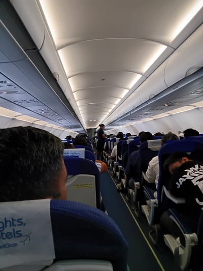The image shows an interior view of an airplane cabin filled with passengers. The seats are arranged in rows, and most are occupied. A person wearing a black hoodie is visible in the foreground, with the headrest of their seat showing the words "flights, hotels," and the website "ndiGo.in." Further down the aisle, a flight attendant in a dark uniform is visible. Overhead compartments line the top of the cabin on both sides, with illuminated panels running along the edges. The general ambiance suggests the cabin is occupied and in transit.