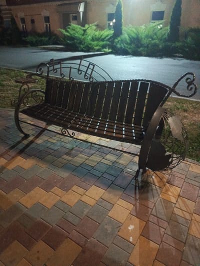 An ornate, dark metal bench is situated on a patterned brick walkway. The bench is designed with intricate scrollwork and features what appear to be musical notes integrated into its backrest. The sides of the bench are constructed with a spherical, cage-like design that resembles a globe, complete with a continental outline on one side. The bench itself has wooden slats for seating. The walkway is paved with rectangular bricks in a herringbone pattern, featuring a mix of reddish-brown, tan, and grey colors. In the background, there is a building with several windows and lush green bushes and trees. The lighting suggests it is nighttime, with artificial lights casting shadows and illuminating the scene. The location is identified as Biaroza, Belarus.