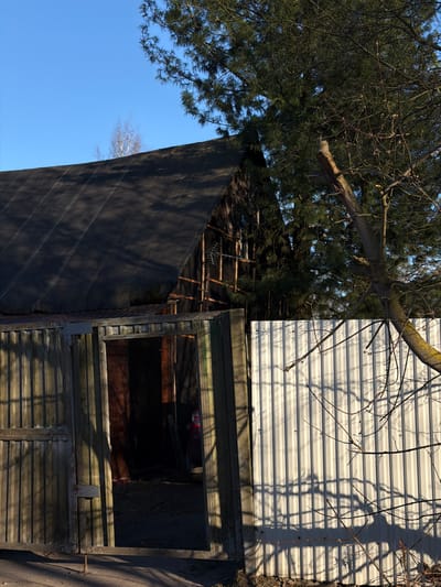 An exterior view shows a ramshackle wooden structure, potentially a shed, with an open window, set against a backdrop of blue sky and tree branches, and what appears to be the exterior siding of a building with horizontal lines. The setting seems to be in a rural or semi-rural area. There is no evidence in the image to confirm this is in Lavriki, Russia.