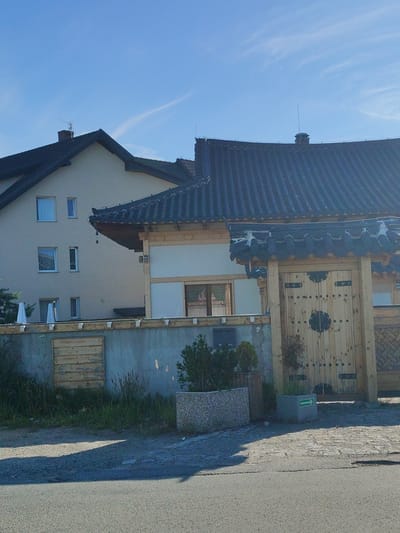 This media file shows a scene in Bielany Wrocławskie, Poland, featuring a building with traditional Korean architectural elements situated next to a more modern house. The foreground is dominated by an asphalt road and a cobblestone pathway. To the left of the image, a light-colored, multi-story house with a dark tiled roof and several windows is visible. To the right, a building with a distinct dark, curved tiled roof, characteristic of traditional Korean design, is prominent. This structure has a large wooden gate with decorative metalwork and round wooden elements. A stone wall separates the modern house from the Korean-style building. Various plants and bushes are present in the garden area. A small green sign with a white arrow pointing to the right is affixed to a concrete planter in front of the wooden gate. The sky is a clear blue with a few wispy white clouds, suggesting it is a sunny day. The lighting indicates it is daytime, likely morning or afternoon. There are no people visible in the frame, and the overall atmosphere is serene.