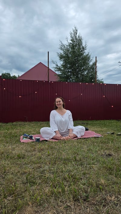 A woman is sitting in a meditative pose on a pink mat in a grassy yard. She is wearing a white outfit. A dark red corrugated metal fence is behind her, decorated with string lights. A red-roofed building is visible behind the fence. The sky is cloudy. The location is Kypoyarovo, Russia.