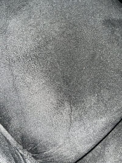 This close-up image depicts a dark, textured fabric surface within the city of Alasia, Nigeria. The material is uniformly charcoal grey to black, exhibiting a dense, short-pile texture consistent with microsuede or a similar upholstery fabric. Variations in light reflectivity across the surface highlight the subtle nap and minor irregularities. A distinct, curved seam or fold is present in the lower right quadrant of the frame, suggesting the fabric is part of an upholstered object or garment. Scattered small, light-colored particulates, consistent with dust or lint, are visible across the surface. No individuals, actions, or specific environmental features beyond the textile itself are discernible.