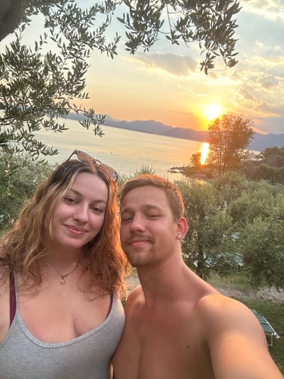 A young couple takes a selfie outdoors at sunset with a lake and mountains in the background. The woman is on the left, facing the camera with a slight smile. She has long, wavy blonde hair and is wearing a grey tank top with a maroon bra underneath. Sunglasses are perched on top of her head. The man is on the right, also facing the camera with a smile. He has short brown hair and is bare-chested. He has an earring in his left ear.
The background shows a body of water, likely Lake Garda, with mountains visible in the distance. The sun is setting on the horizon, casting a warm glow across the sky and water. Olive trees and other greenery surround the couple, suggesting a Mediterranean setting. The location appears to be a viewpoint overlooking the lake.