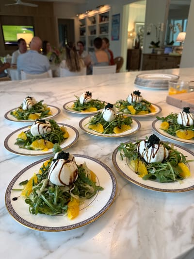In East Naples, United States of America, an indoor scene depicts a gathering of people at a marble-topped counter. Several plates of burrata salads are arranged on the counter, each topped with a ball of burrata cheese, a drizzle of balsamic glaze, arugula, orange slices, and black caviar. In the background, a group of individuals are seated at a dining table, with one person watching a television screen. The environment appears to be a home or restaurant setting, with bookshelves and cabinetry visible in the background.